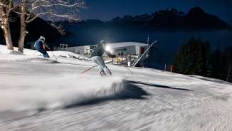 Two skiers look out over the illuminated Rhine Valley while skiing at night on the Pizol