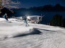 Two skiers look out over the illuminated Rhine Valley while skiing at night on the Pizol