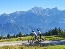 Two people ride downhill on Alpine scooters and the mountains of the Bündner Herrschaft can be seen in the background