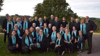 A mixed choir with black clothing and light blue scarves poses outdoors